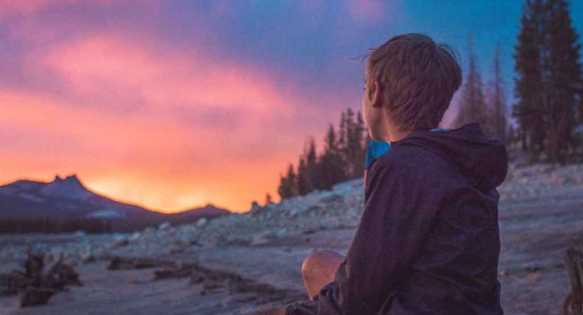 a young person looks away from the camera in the direction of a sunset behind mountains in yosemite